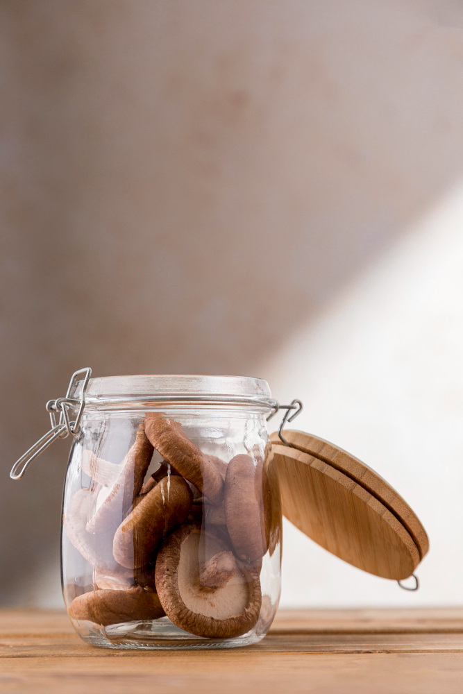 A clear glass jar with a bamboo lid sits on a wooden surface. The jar is filled with dried shiitake mushrooms, and the lid is placed next to the jar.